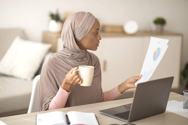 Remote job concept. Focused black Arab woman in hijab reading documents near laptop computer at home