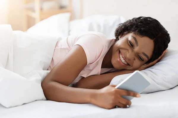 Feliz joven mujer negra usando smartphone en la cama —  Fotos de Stock