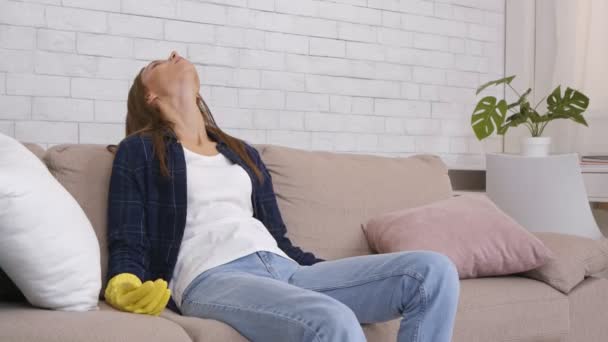 Молодая уставшая женщина в защитных резиновых перчатках сидит на диване, расслабляясь после уборки дома работы на дому — стоковое видео