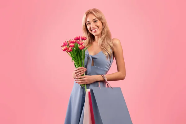 Portrait de jolie jeune femme avec bouquet de tulipes et sacs cadeaux shopping pour les vacances de printemps, fond studio rose — Photo