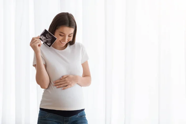 Mujer embarazada feliz sosteniendo ultrasonido escáner de bebé y tocando vientre — Foto de Stock