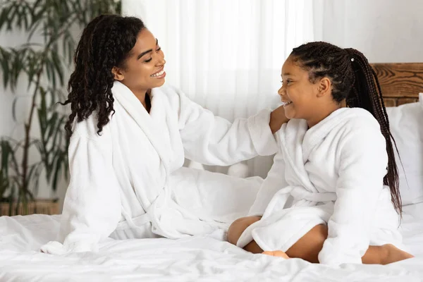 Весела афроамериканська мама і дочка в халатах сидить на ліжку, розслабляючись — стокове фото