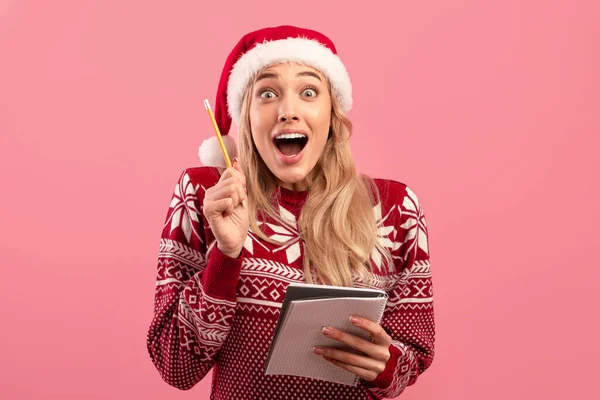 Mujer joven emocionada en jersey de Navidad y sombrero de Santa escritura resoluciones de Año Nuevo en bloc de notas sobre fondo rosa —  Fotos de Stock