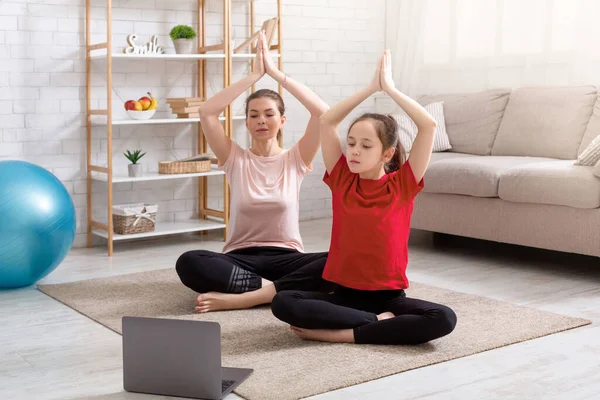 Domestic workout. Young sporty mother and her teen daughter doing yoga to online video course on laptop at home