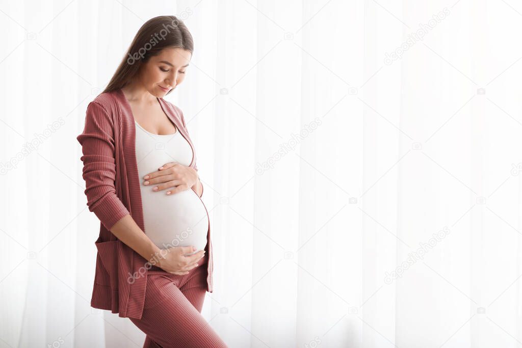 Expecting Baby. Happy Young Pregnant Lady Touching Belly Near Window At Home