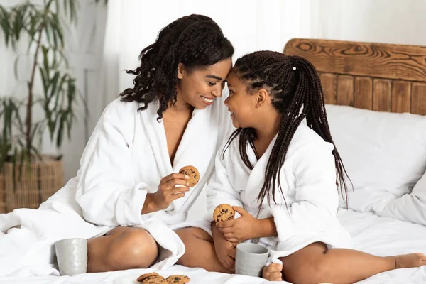 Houden van zwart mam en dochter in badjassen bonding thuis, eten snacks — Stockfoto
