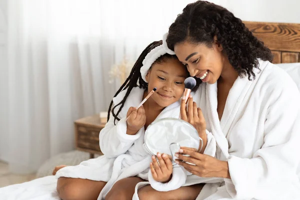 Mãe africana e sua filhinha em roupões de banho fazendo maquiagem na cama — Fotografia de Stock