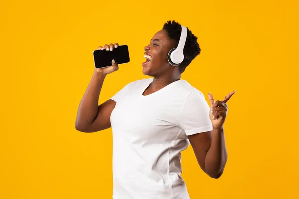 Plus-Size fekete nő fülhallgató éneklés gazdaság Smartphone, sárga háttér — Stock Fotó