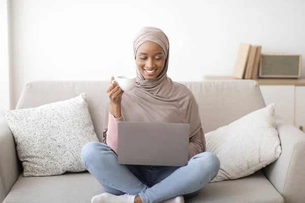 Mulher negra alegre no hijab ter vídeo chat com a família ou amigo no laptop em casa, beber café no sofá acolhedor — Fotografia de Stock