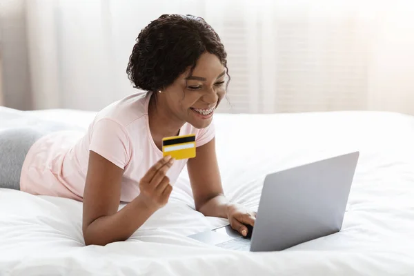 Yatakta kredi kartı ve bilgisayarla alışveriş yapan mutlu bir kadın. — Stok fotoğraf