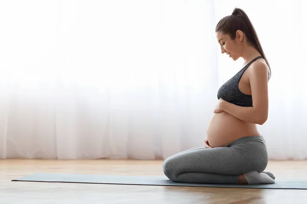 Беременная женщина тренирует йогу дома, сидит на коврике и трогает живот — стоковое фото