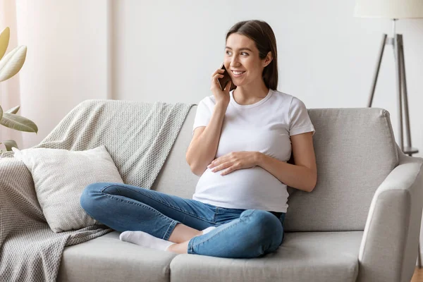 Wanita hamil ceria berbicara di ponsel dan memeluk perutnya di rumah — Stok Foto