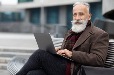Zengin, gri saçlı, yaşlı bir adam ofis binasının yanında oturuyor, dizüstü bilgisayar kullanıyor.