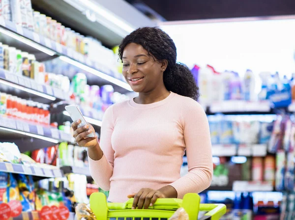 スマートフォンのモバイルショッピングリストアプリを使用して、スーパーマーケットで商品を購入する若い黒人女性 — ストック写真