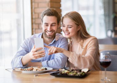 Neşeli milenyum çifti rahat bir kafede romantik bir akşam yemeği sırasında internetten akıllı telefonlarına bakıyorlar.