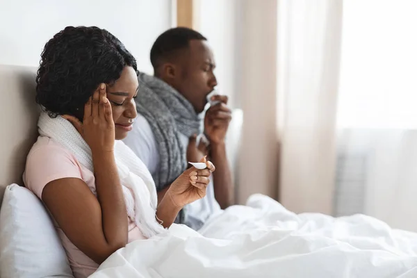 Sjukt svart par som sitter i sängen, mäter feber, nyser näsor — Stockfoto
