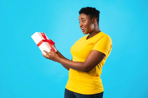 Возбужденный Plus-Size Африканская женщина держит подарок стоя на синем фоне — стоковое фото
