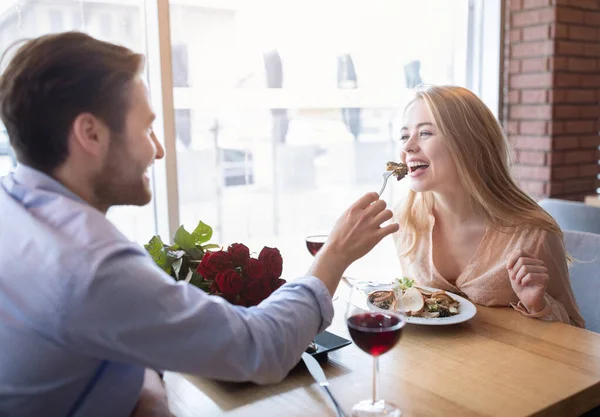 Schönes junges Paar beim romantischen Abendessen mit Blumen und Wein im Café, Millennial Kerl füttert seine Freundin — Stockfoto