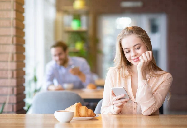 Első ismerősöm. Csinos fiatal nő ellenőrzi az okostelefonját a kávézóban, jóképű fiatal srác néz rá, szabad hely — Stock Fotó