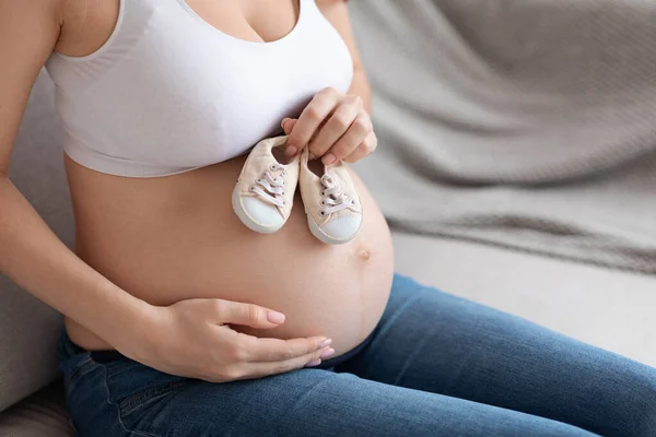 Esperando a Baby. Mujer embarazada irreconocible abrazando el vientre desnudo y sosteniendo zapatos pequeños — Foto de Stock