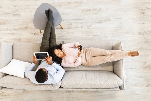 Africano casal americano sentado no sofá, usando tablet e celular — Fotografia de Stock