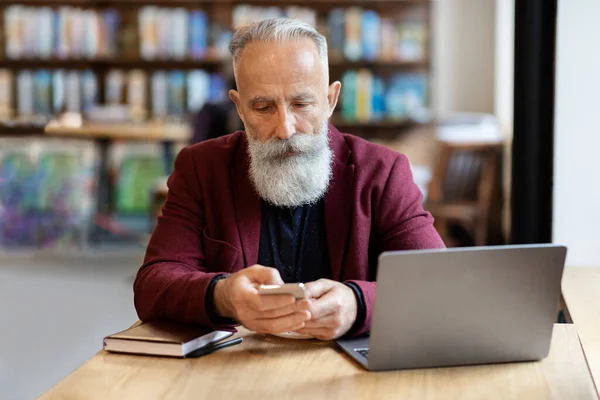 Betagter Geschäftsmann sitzt mit Smartphone vor Laptop — Stockfoto