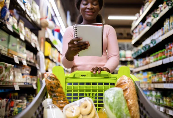 Ung svart kvinna med kundvagn full av produkter som tittar in i hennes livsmedelslista på snabbköpet — Stockfoto