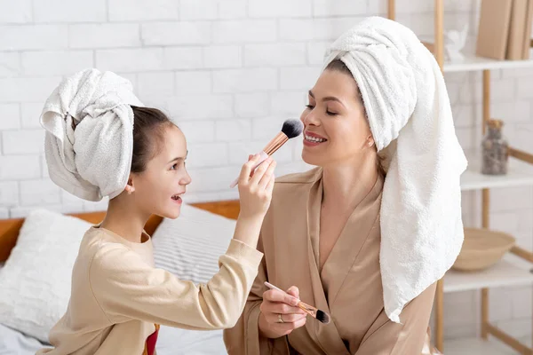 Веселая мама и ее дочь в банных полотенцах, веселятся с макияжем, наносят порошок с кисточками на кровать дома — стоковое фото