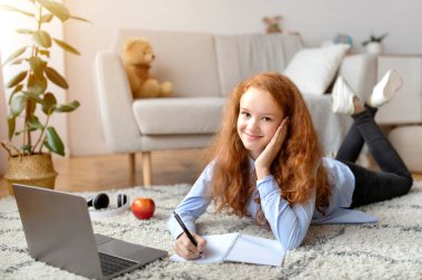 Kız yerde yatıyor, dizüstü bilgisayar kullanıyor, deftere yazıyor.