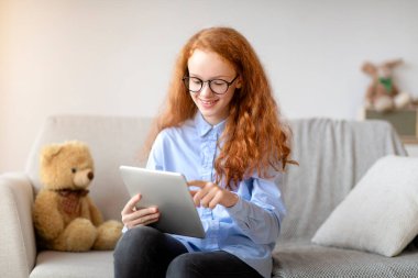 Dijital tablet kullanan gözlüklü kız, kanepede oturuyor.