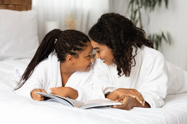 Αφρικανή μαμά και κόρη χαλαρώνουν στο κρεβάτι με μπουρνούζια και διαβάζοντας βιβλίο — Φωτογραφία Αρχείου