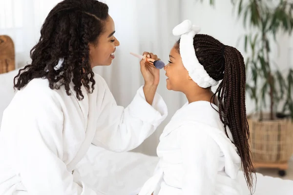 Amar a mãe africana e a menina em roupões de banho aplicando maquiagem em casa — Fotografia de Stock