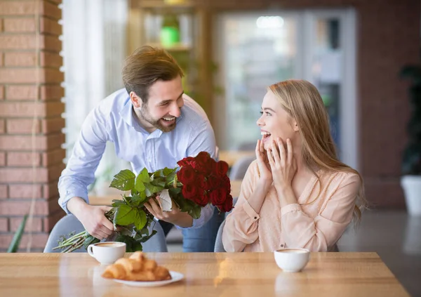 Красивий молодий хлопець представляє букет квітів своїй збудженій дівчині в кафе, що робить її особливим сюрпризом — стокове фото