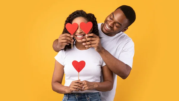 Black Husband Covering Wifes Eyes With Hearts Over Yellow Background — Stok Foto