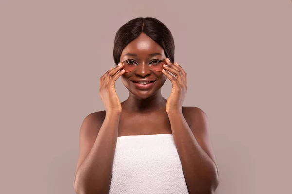 Tratamento da pele dos olhos. Mulher afro-americana sorrindo aplica manchas douradas sob os olhos, tem toalha branca na cabeça, assistindo no espelho, no spa — Fotografia de Stock