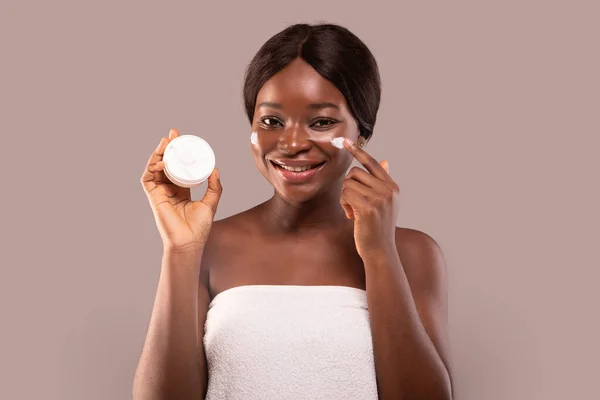 Cuidado com a pele. Sorrindo mulher africana em toalha aplicando creme hidratante no rosto — Fotografia de Stock