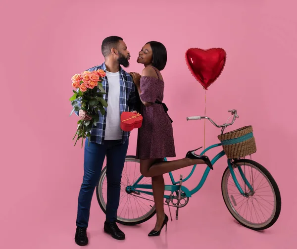 Pełna długość portret pięknej czarnej pary z różami, Walentynki prezent i całowanie rowerów na różowym tle studio — Zdjęcie stockowe