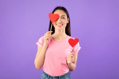 Gülümseyen kadın iki kalp tutuyor, bir gözünü kapatıyor.