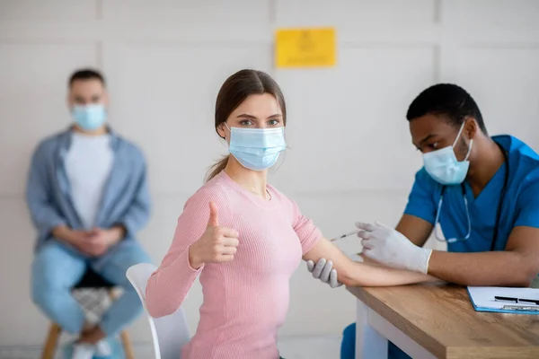 Mujer joven en máscara médica aprobando la vacunación con covid-19, mostrando un gesto de pulgar hacia arriba en la clínica —  Fotos de Stock
