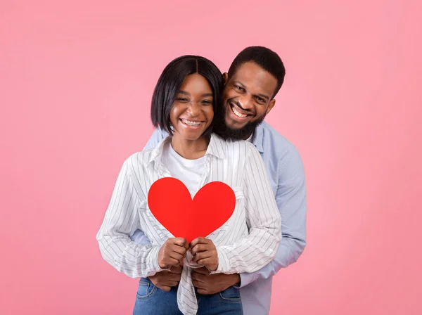 Valentines Day holiday. Portrait of positive black couple holding red paper heart and hugging on pink background — Stock Photo, Image