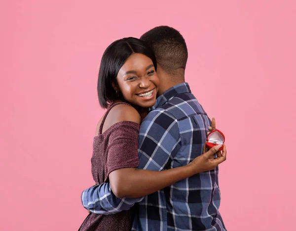 Feliz mujer negra abrazando a su prometido y sosteniendo el anillo de compromiso en el fondo rosa del estudio — Foto de Stock