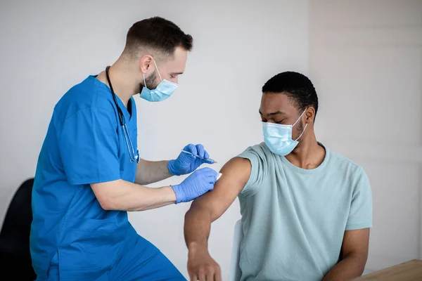 Médico con mascarilla facial que administra covid-19 vacuna inyectable al paciente varón negro durante la campaña de vacunación en la clínica —  Fotos de Stock