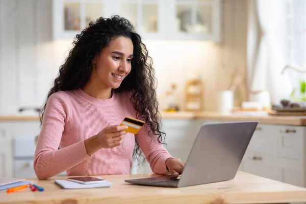 Compras en línea. Sonriente morena mujer usando tarjeta de crédito y portátil en la cocina — Foto de Stock