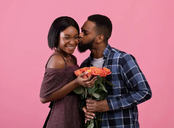 Afectuoso chico negro dando a su novia ramo de flores para el día de San Valentín y besándola sobre fondo rosa — Foto de Stock