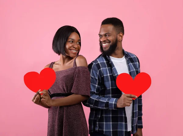 Hari Valentine. Tampan hitam pria dan pacarnya memegang hati merah dan melihat satu sama lain di latar belakang merah muda — Stok Foto