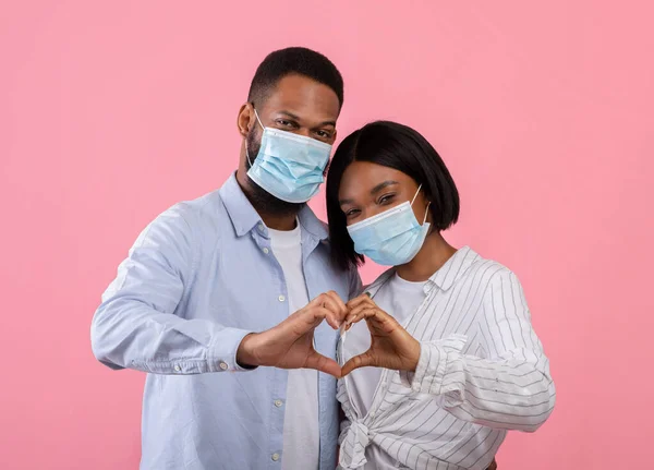 Valentines Day during covid-19 outbreak. Black couple in face masks making heart with their hands on pink background — Stock Photo, Image