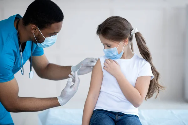 Joven médico negro haciendo la inyección de la vacuna covid-19 a una niña en mascarilla facial en el hospital — Foto de Stock