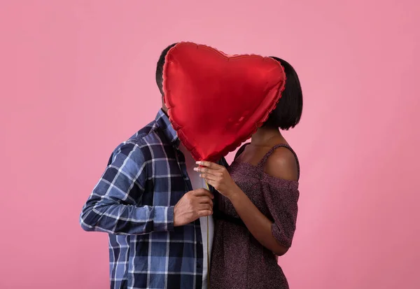 Романтична чорна пара цілується за кулею серцевої форми на День святого Валентина над рожевим фоном студії, вид збоку — стокове фото