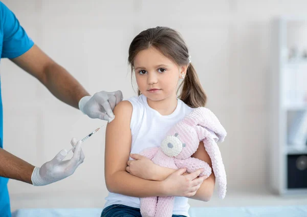 Jolie petite fille avec un lapin jouet recevant une injection de vaccin covid-19 à la clinique. Immunisation contre le virus mondial — Photo