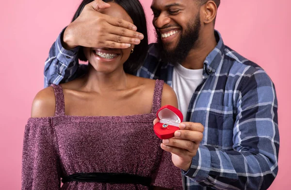 Szczęśliwy Afroamerykanin zakrywający oczy narzeczonych, trzymając pudełko z pierścionkiem zaręczynowym na różowym tle — Zdjęcie stockowe